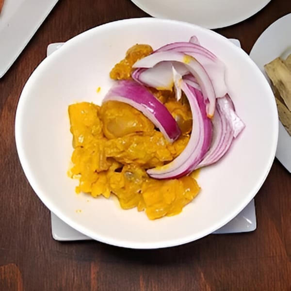 A white bowl of BUKA New York's African food, featuring yellow stewed meat and red onion slices.