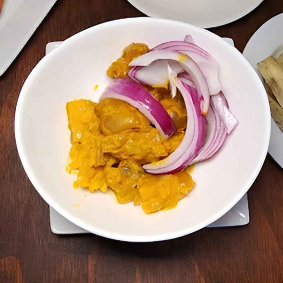 A white bowl of BUKA New York's African food, featuring yellow stewed meat and red onion slices.